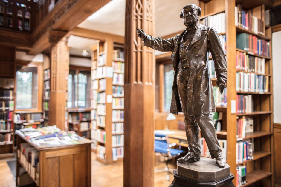 A bronze statue of a distinguished man in a long coat and trousers, standing on a black pedestal, with one arm extended as if addressing an audience, located inside a warmly lit library with wooden bo