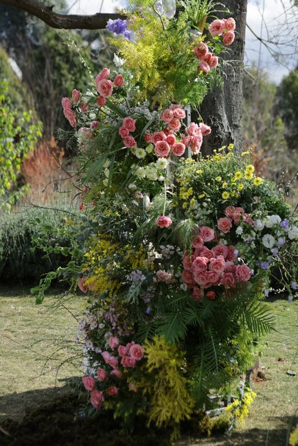A tall floral arrangement standing outdoors against a natural background with trees and grass, featuring a variety of fresh flowers. The bouquet includes clusters of pink roses and carnations, complem