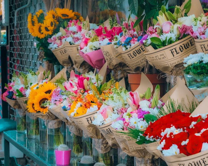 A colorful display of fresh flower bouquets arranged on a glass shelf at a floristry shop, featuring vibrant sunflowers, pink and white lilies, orange and pink blossoms, and red roses, each wrapped in