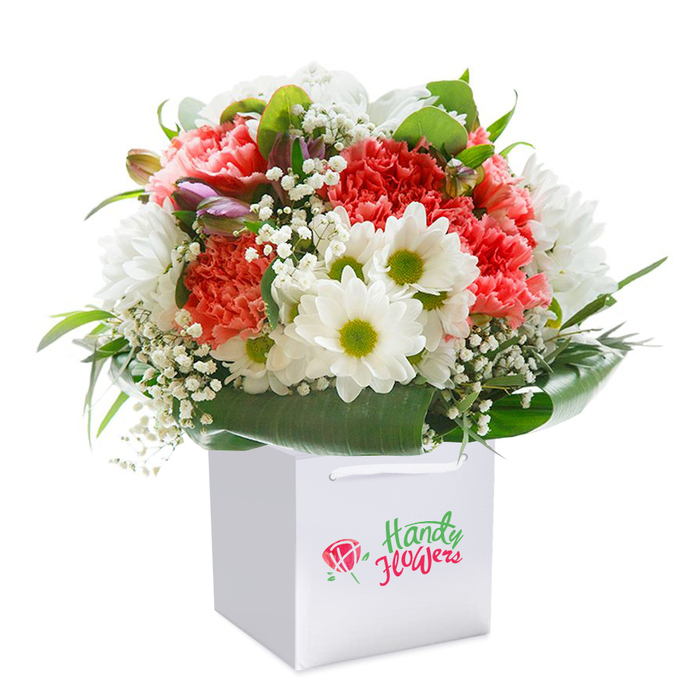 This eye-catching floral arrangement, meticulously assembled by a Willesden florist, is elegantly presented upright in a sleek white square gift bag with supporting ribbon handles. The bouquet boasts a full, rounded composition, highlighted by lavish coral and peach-toned carnations recognized for their lush, ruffled petals. Bright white daisy-style chrysanthemums with vibrant lime-green centers add cheerful contrast throughout, their simplicity evoking community gardens found off Harlesden Road. Sprays of delicate gypsophila weave a gentle, cloudlike texture among the dominant blooms, softening the overall structure. Nestled discreetly within the bouquet, closed buds showing hints of purple and pink add both mystery and harmony, suggesting blossoming moments yet to come. Broad, deep green leaves and glossy foliage encircle the floral core, providing a stately base and contributing to the arrangement's balanced, abundant silhouette. Against a stark white studio background, the colours appear crisp and lively, and the clean presentation reflects the professionalism of Willesden's flower deliveries. Every stem is positioned with intention, suitable for gifting on birthdays, anniversaries, or any occasion that calls for a sincere, elegant touch within the NW2 community. The image is designed to evoke both the freshness and thoughtful care that define local Willesden floristry.