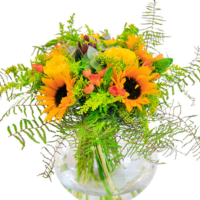 The atmosphere of Willesden comes alive in this exuberant yellow and orange floral display, expertly arranged to reflect the energy of bustling marketplaces and tranquil corners of King Edward VII Park. Radiant sunflowers, with their broad russet-brown centers and luminous petals, stand tall, commanding the eye's attention and conjuring up the height of summer's bounty. Around them, plump garden roses unfurl in spirals of creamy yellow, their soft petals almost glowing with warmth. Scattered throughout are clusters of bright red Hypericum berries, their small, polished forms offering delightful contrast and recalling the cheerful colors of local shopfronts. The arrangement is lavishly padded with variegated greenery: wispy fern fronds add delicate motion, while slender spikes of green and sprays of goldenrod mirror the textures of Willesden's verdant parks. Soft, matte eucalyptus leaves peek between the blooms, lending an aromatic freshness and a gentle blue-green highlight. All these elements are anchored in a clear, spherical vase that allows the green stems to be fully visible, as if growing straight from a peaceful backyard garden. Carefully crafted for joyous occasions, from anniversaries to warm reunions, this bouquet brings the luminous character of Willesden into every room -- its scent, colors, and textures embracing you with each glance and breath.