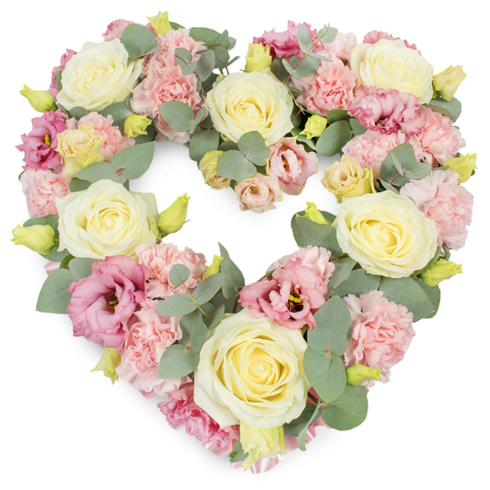 In this image, a lush, heart-shaped sympathy wreath stands out clearly against a plain white background, allowing every pastel shade and petal texture to be seen in detail. The heart is complete and symmetrical, its interior entirely filled with blooms so that the overall look is dense, luxurious, and deeply considered. The most prominent flowers are large cream and pale lemon-yellow roses, each bloom carefully spaced to act as visual anchors around the heart. Their petals form soft spirals, transitioning from tighter centres to gently unfurling edges that catch subtle highlights, suggesting grace and enduring love. Between and around these roses, numerous pastel pink carnations create a billowy, almost cloud-like layer. Their many ruffled petals overlap and mingle, bringing a feathery softness that contrasts gently with the smoother rose heads. Pockets of richer colour appear in the form of magenta to mauve-pink lisianthus, their more open, cup-shaped flowers adding depth and drawing the eye deeper into the arrangement. Tiny, not-yet-open yellow-green buds punctuate the design, like quiet reminders of renewal and the ongoing cycle of life. Winding through the flowers are cool, silver-green eucalyptus stems, their rounded leaves and subtle fragrance evoking calm walks through the greener corners of Willesden, perhaps near Gladstone Park. The greenery outlines and supports the heart, giving the wreath a natural, slightly woodland feel while keeping the overall mood refined and respectful. This tribute feels especially suited to funerals or memorial services held in and around Willesden, conveying sympathy, remembrance, and a gentle, comforting presence to family and friends gathered to say goodbye.
