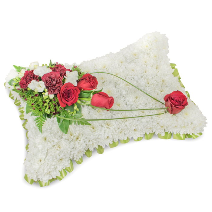 A thoughtful, classic pillow of remembrance, composed with the restraint and care of an experienced Willesden florist at Flowers Willesden. The rectangular cushion is completely cloaked in hundreds of small, immaculate white chrysanthemums whose densely packed faces create a soft, tactile surface that reads as comfort and dignity. Along the perimeter a pale green satin ribbon or narrow foliage trim peeks through the blooms, forming a gentle lime-toned outline that neatly finishes the shape. On the upper left a rich floral spray commands attention: several deep red roses in varying stages of bloom sit beside dark burgundy carnations, their layered petals offering a luxurious contrast against the white field. Interwoven are small white freesias and fine spray chrysanthemums that introduce delicate highlights and variation. Surrounding the cluster, glossy green leaves and feathery fern fronds give the arrangement body and a natural silhouette. From this focal cluster, three sinuous stems of bright green bear-grass or decorative grass curve in an elegant arc across the cushion, visually connecting to a single prominent red rose placed in the lower right - a poetic finishing touch that suggests devoted remembrance. The piece is shown in soft, even light that emphasizes texture and tone; the subtle fragrance is clean, green and reassuring. Designed for sympathy and memorial settings, this cushion carries a message of lasting love and respect and is appropriate for tributes at local chapels, graveside services near Kensal Green Cemetery, or intimate wakes around Harlesden and Willesden High Road.