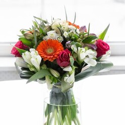 A celebration of colour and craftsmanship unfolds in this lavish Willesden bouquet, lovingly presented in a tall, clear glass vase. The composition revolves around the dynamic interplay between vibrant orange and creamy peach gerbera daisies, each petal catching flecks of light much like the dappled sunshine that spills across Willesden's charming side streets. Nestled beside them, sumptuous magenta roses stand out for their rich tones and tightly-wound petals, symbols of refined romance and exuberance. White alstroemeria and sprays of petite, star-shaped white flowers wind their way through the arrangement, bringing texture and balance. Pale pink accents and tiny white florets create further layers of depth, inviting lingering appreciation. Lush green foliage, from feathery sprigs to substantial, glossy broad leaves, encircle the bouquet; the aspidistra leaves are curved with the care of a seasoned florist, wrapping the blooms in a protective green embrace that grounds the colourful display. Through the crystal vase, vibrant stems are clearly visible-a reminder of the bouquet's fresh, garden-like character. Bathed in soft, natural light, this arrangement conveys a spirit of renewal and generosity, making it perfect for gatherings along the High Road, as a centrepiece in a Willesden home, or as a heartfelt gesture to mark new beginnings within the local community.